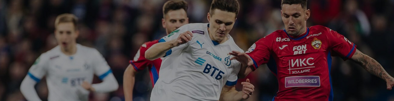 На изображении два футболиста соперничают на поле во время матча. Один в белой форме, другой в красно-синей. Они сосредоточены на мяче, а на заднем плане виден еще один игрок. На форме логотипы спонсоров