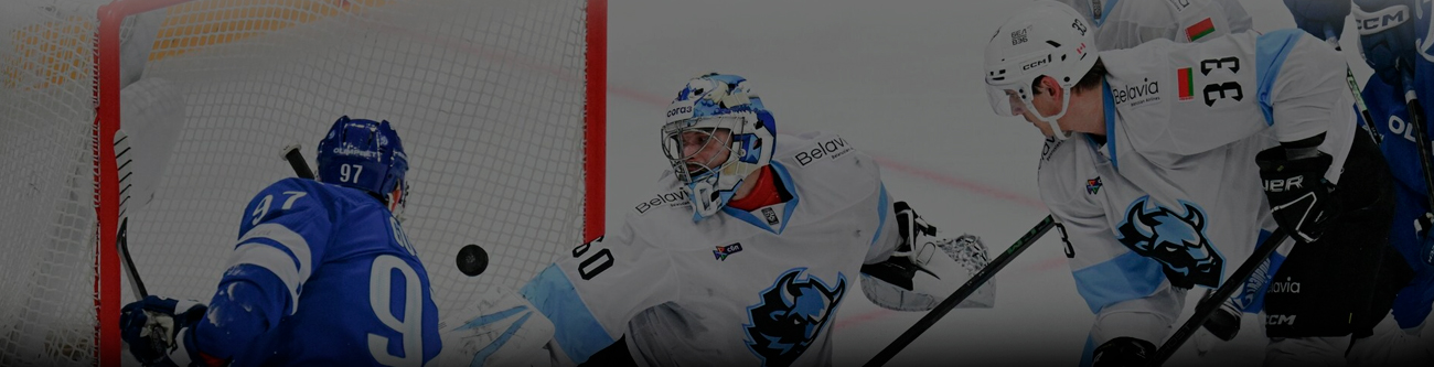 На изображении момент хоккейного матча: игрок в синей форме атакует ворота соперника, защищаемые вратарем и защитниками в бело-голубой форме. Напряженная борьба разворачивается рядом с вратарской зоной