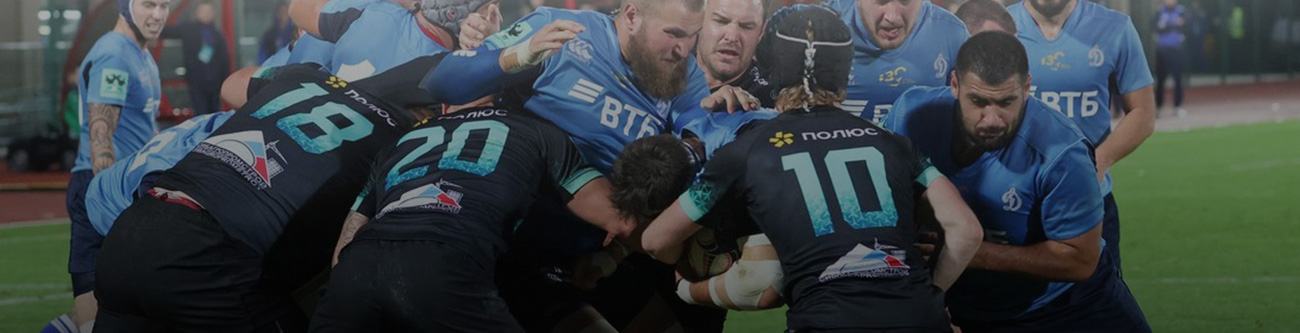 На изображении запечатлен момент игры в регби. Две команды в синих и черных формах участвуют в схватке за мяч на зеленом поле. Игроки напряжены и сосредоточены, демонстрируя командную работу и решимость