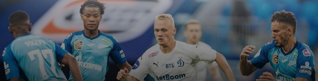 На изображении футболисты в синих и белых формах соревнуются на поле. Три игрока в форме одного цвета, один в другом. На заднем плане трибуны стадиона и размытый рекламный баннер