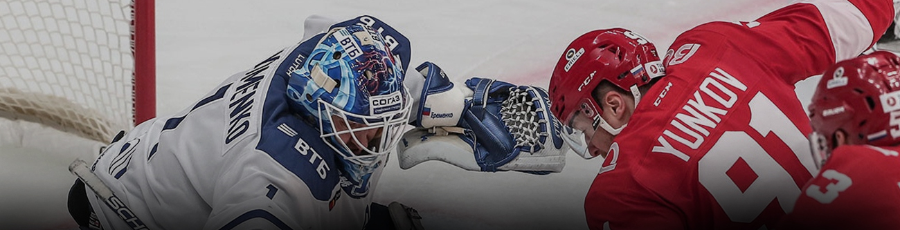 Вратарь в синей форме тянется за шайбой, пытаясь остановить её, в то время как хоккеист в красном с соревновательным запалом направляет шайбу в сторону ворот. На заднем плане видны ворота