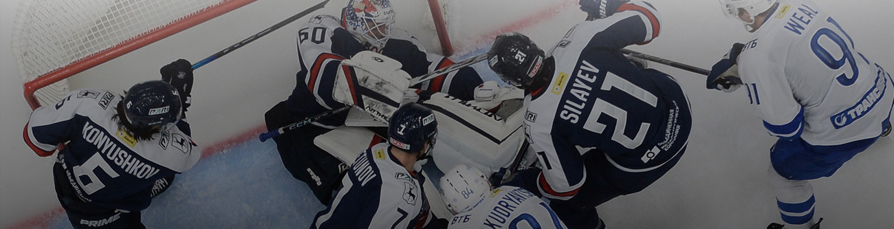 На изображении хоккейный матч, игроки в тёмной форме блокируют вратаря в белой форме, в борьбе за шайбу перед воротами. Видны разные номера и фамилии на спинах хоккеистов. Игра проходит на ледовом поле