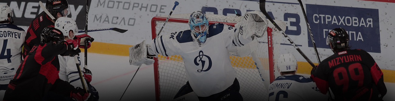 На изображении хоккейный матч: вратарь в белой форме защищает ворота, вокруг него игроки в чёрной и красной форме. Справа виден игрок с номером 99. На заднем плане рекламные вывески на борту