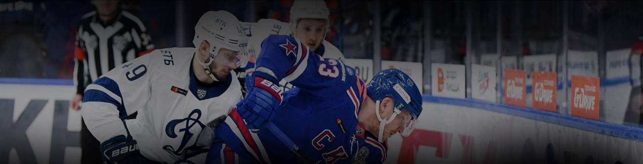 Хоккейный матч: игрок в бело-синей форме с цифрой 9 на рукаве атакует игрока в синей форме. На заднем плане видны ещё один игрок и судья в полосатой форме. На бортике расположены логотипы и рекламные баннеры