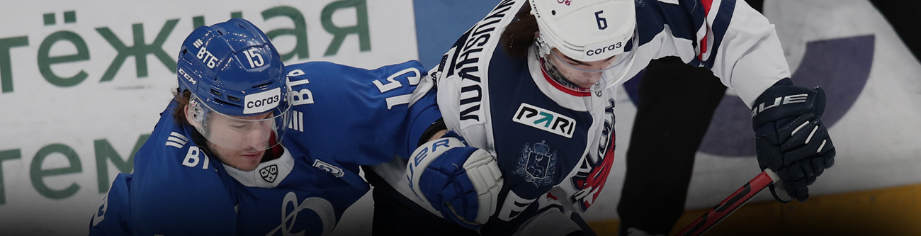 На изображении два хоккеиста соперничают за шайбу на льду. Один в синей форме, другой в белой. Обе команды борются за контроль, демонстрируя динамичность и напряженность игры