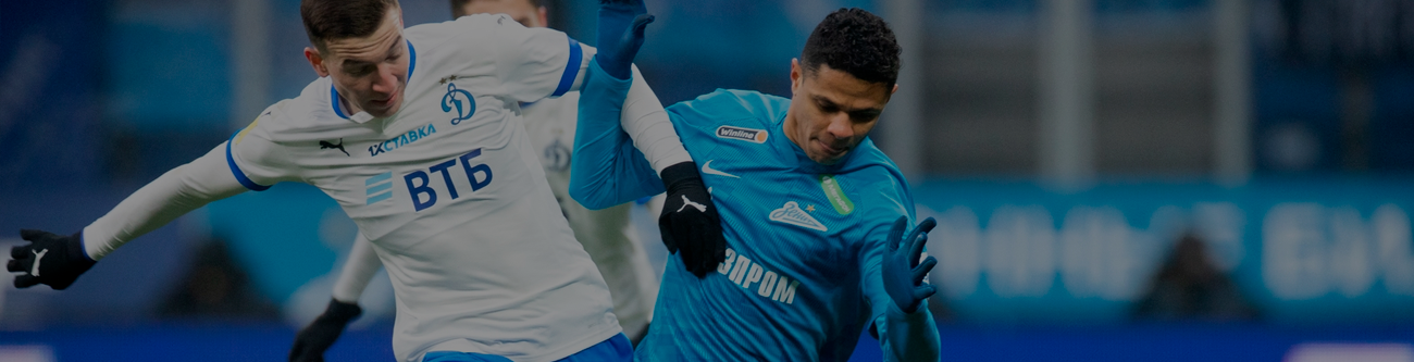 Два футболиста в борьбе за мяч на поле во время игры. Один в бело-синей форме команды, другой — в форме сине-зелёного оттенка. На заднем плане видны размытые зрители и трибуны